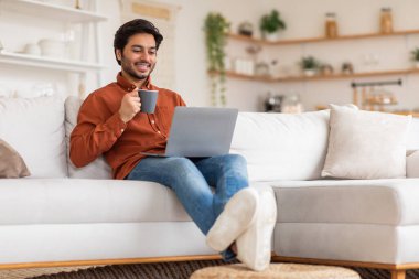 Beyaz bir koltukta oturan bir adam elinde bir fincan kahve tutarken dizüstü bilgisayarını rahatça kullanıyor. Sıradan bir turuncu gömlek, kot pantolon ve beyaz spor ayakkabı giyiyor.