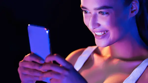 stock image A young woman is seated, holding a smart phone in her hands and looking at the screen intently in a dark room at night. The glow from the phone illuminates her face as she interacts with the device.