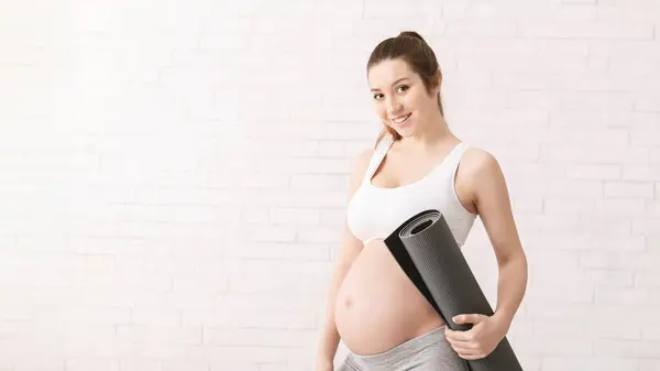 stock image Sporty pregnant woman is standing while holding a yoga mat in her hands. She is visibly expecting and appears to be preparing for a yoga session, copy space