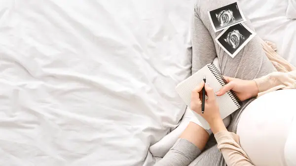 stock image A pregnant woman is seated comfortably in bed, writing in a journal. Beside her are ultrasound images of her unborn baby, top view, copy space