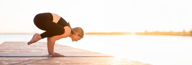 Karga pozisyonunda duran sportif orta yaşlı kadın dışarıda yoga yaparken Nehir kenarındaki tahta iskelede, Dengelenmiş Vücut ağırlığını dirsekleriyle dengeleyen esnek kadın, dış antremanlardan keyif alan, Kopya Boşluğu