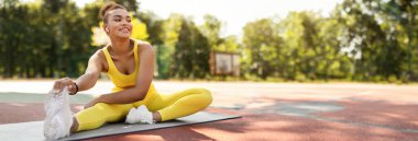 Spor Yaşam Tarzı Konsepti. Sarı bluzlu, sırtüstü uzanan beyaz spor ayakkabılı, yoga minderinin üzerinde oturan, ayaklarına dokunan, uzayı kopyalayan, gülümseyen Afrikalı Amerikalı kadın.