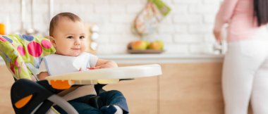 Bir bebek parlak, modern bir mutfakta renkli bir yüksek sandalyede rahatça oturuyor. Arka planda, anne tezgahta yemek hazırlarken görülüyor, bu da tipik bir sabah rutinini gösteriyor..
