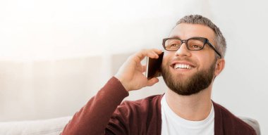 Gözlüklü bir adam cep telefonuyla bir konuşma yapar ve kulağına tutar. Diğer tarafla iletişim kurarken odaklanmış ve dikkatli görünüyor..