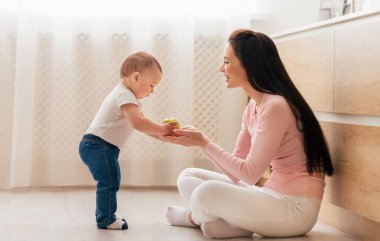 Bir anne yerde oturuyor, bebeğiyle konuşurken gülümsüyor, bebek ayakta duruyor ve ona iyi aydınlatılmış bir oturma odasında oyuncak veriyor..