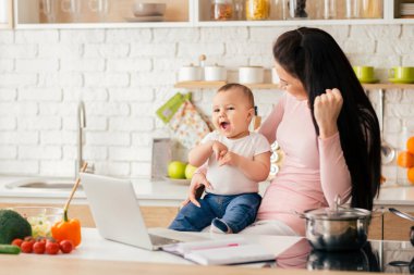 Bir anne ve bebek modern bir mutfakta neşeli bir anı paylaşır. Anne hem gülümseyen hem de meşgul bir şekilde dizüstü bilgisayara bakarken bebek tezgahta oturur..