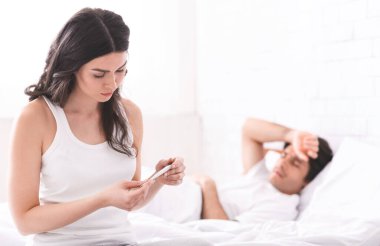 Bir kadın bir yatağın kenarında oturur, termometreye bakar, bir erkek de arkasında yatar. Endişeli görünüyor..
