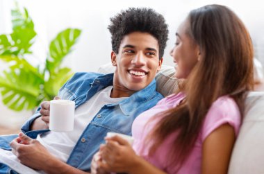 Siyah genç çift bir kanepede oturup sohbet ederken bir fincan kahveden hoşlanırlar. İkisi de mutlu ve rahat görünüyor.