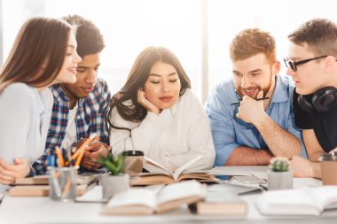 Kütüphane masasındaki çok ırklı öğrenciler öğrenmelerini geliştirmek için kitapları kullanıyorlar. Grupların dinamik etkileşimi modern, kapsayıcı bir eğitim ortamını somutlaştırır.