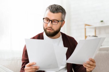 Bir adam kanepeye oturmuş elinde bir yığın kağıt tutuyordu. Belgelere odaklanmış görünüyor, muhtemelen onları okuyor ya da gözden geçiriyor..