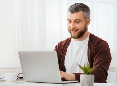 Bir adam dizüstü bilgisayarın önünde oturuyor, ciddi bir ifadeyle ekrana odaklanıyor. Klavyeye yazı yazıyor ve çalışıyor ya da dikkatle çalışıyor gibi görünüyor..
