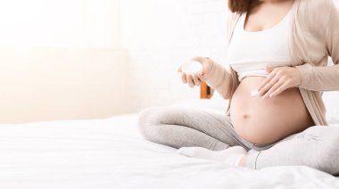 Hamile bir kadının göbeğine nemlendirici krem sürerken, yatakta otururken, boş bir panorama yaparken.