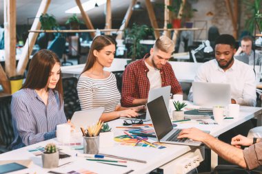 Bir grup genç profesyonel, modern, açık ofis ortamında doğal ışık, dizüstü bilgisayarlar ve tasarım materyalleriyle birlikte çalışıyorlar..