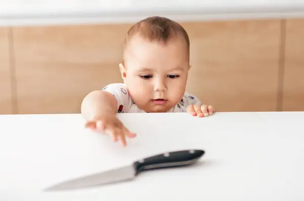 Stock image A curious baby, dressed in a white outfit with colorful patterns, reaches for a knife placed on a kitchen counter. The background features wooden cabinets and a light-colored wall