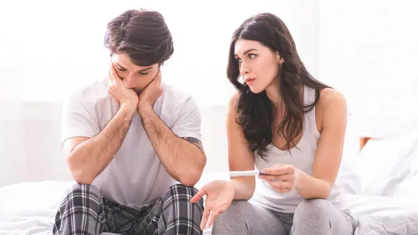 Stock image A woman is sitting on a bed with her arm extended, holding a pregnancy test and looking at a man sitting next to her. The man has his head in his hands, looking down at his lap.