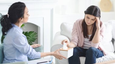 Bir danışman, parlak, rahat bir odada terapi seansı sırasında duygusal ve sıkıntılı görünen genç bir kadına bir kutu mendil sunuyor..