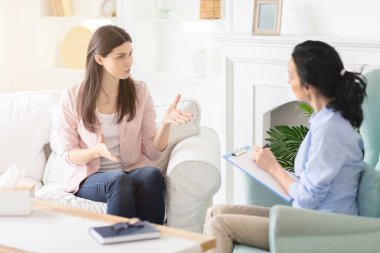 İki kadın iyi aydınlatılmış bir oturma odasında bir kanepede oturuyor. Ciddi bir sohbete girişmişler gibi görünüyor. Bir kadın panoya not alıyor..