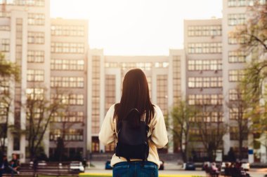 Bir kadın yüksek bir binanın önünde duruyor, etkileyici yüksekliğine bakıyor. Binanın şaşaalı mimarisi ve cam cephesi gökyüzüne karşı kontrasttır.