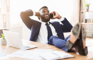 Ofiste mola verirken telefonla konuşan neşeli bir Afrikalı menajer sandalyeye yaslanıp bacakları masaya koyuyor ve fotokopi alanı olan bir panorama yaratıyor.