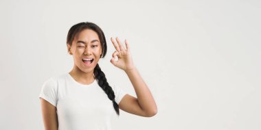 Genç siyah bir kız eliyle iyi bir jest yaparken resmediliyor. Yüzünde tarafsız bir ifade var ve sıradan bir şekilde hareket ediyor gibi görünüyor..