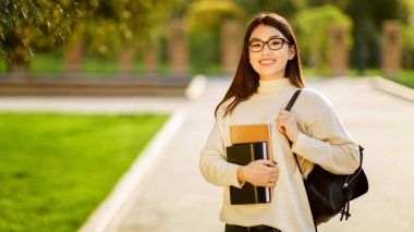 Gözlüklü Asyalı genç bir kadın tüm sabah bir üniversite kampüsünde elinde kitaplarla ve sırt çantasıyla yürüyor. Açık renkli bir kazak giyiyor ve gülümsüyor.