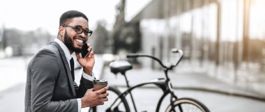 Gülümseyen Afro-Amerikalı işadamı elinde kahve fincanı ve telefonla, şehir ortamında bir bisikletin yanında oturuyor, web-afişi