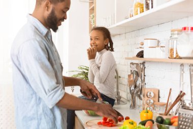 Siyah adam, tatlı kızıyla yemek yapıyor, sebze kesiyor, kurabiye yiyor ve gülümsüyor, yan manzara, kopyalama alanı.