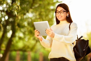 Gözlük takan ve kablosuz kulaklık takan Asyalı genç bir kadın güneşli bir parkta dururken tablet kullanıyor. Sırt çantası var ve gülümsüyor. Açık havanın tadını çıkarıyor..