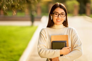 Asian young woman wearing glasses and earbuds is seen walking on a college campus, holding several books and smiling warmly during a sunny day. clipart