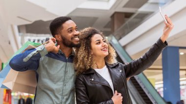 Afro-Amerikalı bir erkek ve kadın hareketli bir alışveriş merkezinde duruyorlar, ellerinde bir cep telefonu ile selfie çekiyorlar, gülümsüyorlar ve dükkan ve müşterilerin önünde poz veriyorlar..