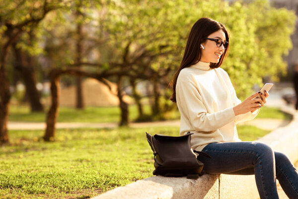 Азиатка сидит на скамейке, поглощённая своим мобильным телефоном. Она выглядит сосредоточенной на экране, отталкиваясь пальцами от текста или прокручивая контент.