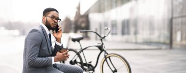 Profesyonel bir siyah adam kahvesini yudumluyor ve telefonuyla konuşuyor, bisikletinin yanında oturuyor. Gündelik ama verimli bir ortam, modern bir şehir ortamında birden fazla iş yapma anını yakalıyor..