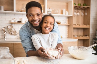 Afro-Amerikan bir baba ve genç kızı kendi mutfağında yemek pişirirken aile ve babalığı simgeliyor.