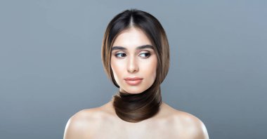 Uzun, pürüzsüz kahverengi saçlı genç bir kadın zarif bir şekilde boynuna dolanmış ve düz gri bir fonda poz veriyor. Yüzü sakin ve düşünceli..