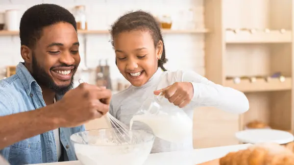 stock image Smiling african american father looks on as his daughter excitedly pours milk into a mix, capturing family involvement in the kitchen