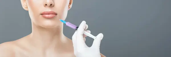 stock image A professional in a clinic setting administers a cosmetic injection near a woman lips, aiming for lip enhancement. The practitioner wears a white glove and wields a syringe, cropped, copy space