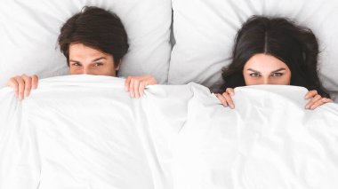 Bir adam ve bir kadın yatakta yatıyor, ikisi de kısmen beyaz bir battaniyenin altında. İkisi de kameraya şakacı, muzip bir ifadeyle bakıyor..