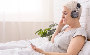 Son sınıf öğrencisi kadın telefonuyla müzik dinlerken yatakta rahat bir şekilde yatıyor. Cihazdan gelen sese odaklanmış ve rahatlamış görünüyor.