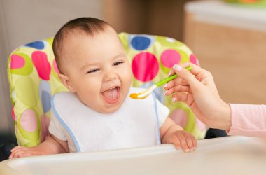 Neşeli bir erkek bebek renkli bir sandalyede oturur, bir bakıcı olarak hevesle ağızlarını açar parlak, neşeli bir mutfakta bir kaşık bebek mamasıyla onları besler..