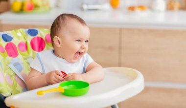 Bir bebek önünde yeşil bir kase ve sarı bir kaşıkla yüksek bir sandalyede otururken neşeyle gülüyor. Ayarlar mutfak gibi görünüyor, çeşitli mutfak malzemeleriyle.