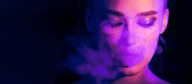 Siyah arka planda parlak bir neon ışığın önünde sigara içen bir kadın görülüyor. Etrafında uçuşan dumanla sigarasına odaklanmış görünüyor.