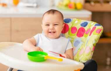 Mutlu bir bebek yüksek bir sandalyede oturur, gülümser ve renkli oyuncaklarla oynar. Modern mutfak ortamı parlak ve neşeli bir atmosfer sağlıyor..