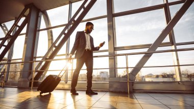 Bavullu profesyonel bir zenci havaalanında telefonuyla bekliyor. Atmosfer dinamik ve modern, iş seyahatini ve bağlantıyı yansıtıyor, güneş parlıyor.