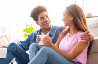 Afro-Amerikalı genç bir erkek ve kız birlikte bir koltuğun yanında oturuyor, kahve içiyor ve gülüyorlar. Her ikisi de günlük olarak kot ve t-shirt giyiyorlar..