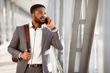 Havaalanı terminalinin dışında bavullu siyah bir adam telefonunu kullanıyor. Sahne canlı ve profesyonelce, iş seyahatinin çoklu görev doğasını vurguluyor..