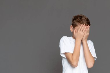 Gri bir arkaplanda, genç bir çocuk perişan ya da şaşkın bir şekilde yüzünü evrensel bir insan ifadesiyle örtüyor.