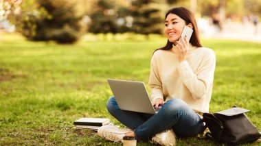 Genç bir kadın parkta çimlerin üzerinde oturuyor, dizüstü bilgisayarı üzerinde çalışırken telefonla konuşuyor. Kitaplar ve çanta yakınlarda, okuduğunu gösteriyor..