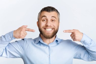 Mavi gömlek giyen bir adam elleriyle işaret ediyor, beyaz dişlerini gösteriyor. Vücut dili vasıtasıyla iletişim kurarken yüz ifadesi odaklanmış görünüyor..