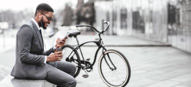 Afrikalı Amerikalı bir adam bisikletinin yanında otururken telefonunu kullanıyor. Sahne açık havada, bir şehir manzarasında iş ve eğlence karışımı, web-afişi sağlıyor.