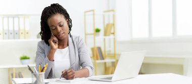 Profesyonel kıyafetli genç Afrikalı Amerikalı iş kadını ofisinde dizüstü bilgisayar kullanıyor, telefon görüşmesi yapıyor, notlar alıyor.